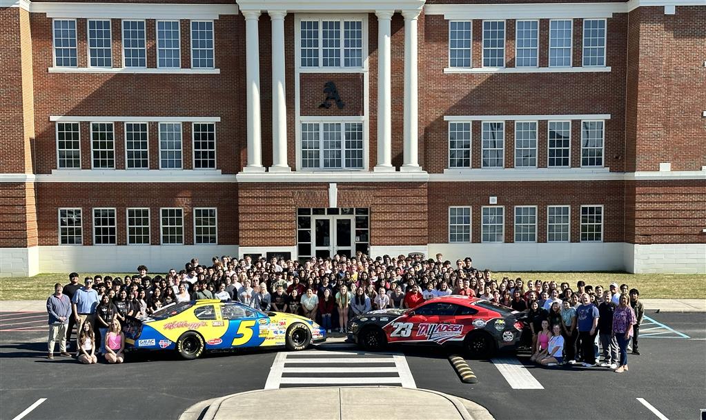 Several students standing in front of school behind two race cars